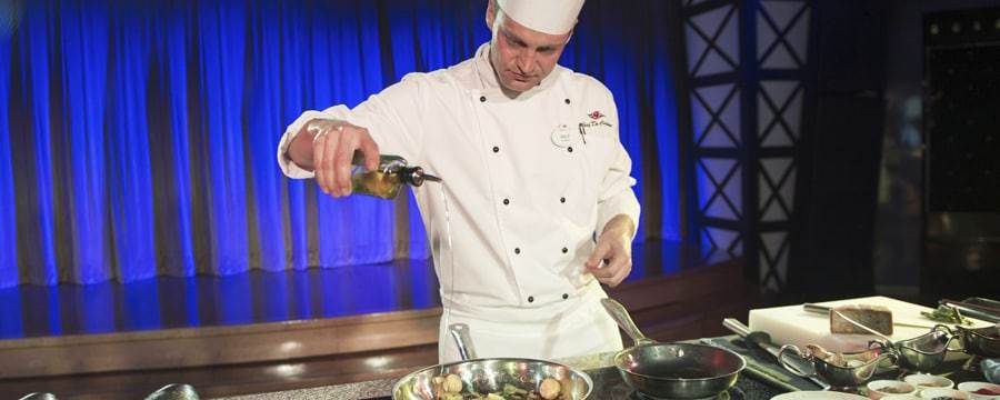 A chef in a demonstration kitchen pours oil into a pan while sautéing scallops with vegetables